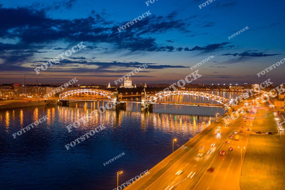 На фотографии изображён Большеохтинский мост, соединяющий берега реки Невы. Подсветка моста отражается в водах Невы, создавая живописную симметрию. На горизонте виден величественный Смольный собор с белыми куполами. Справа проходит оживлённая магистраль, подчёркивающая ритм вечернего города. Снимок выполнен с высоты и передаёт красоту Петербурга в час заката.